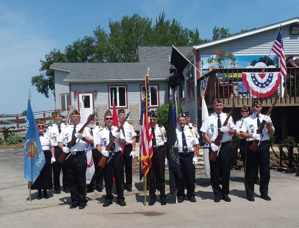 A group of VFW veterans holding flags and rifles in their honor guard uniforms standing in front of a buliding.